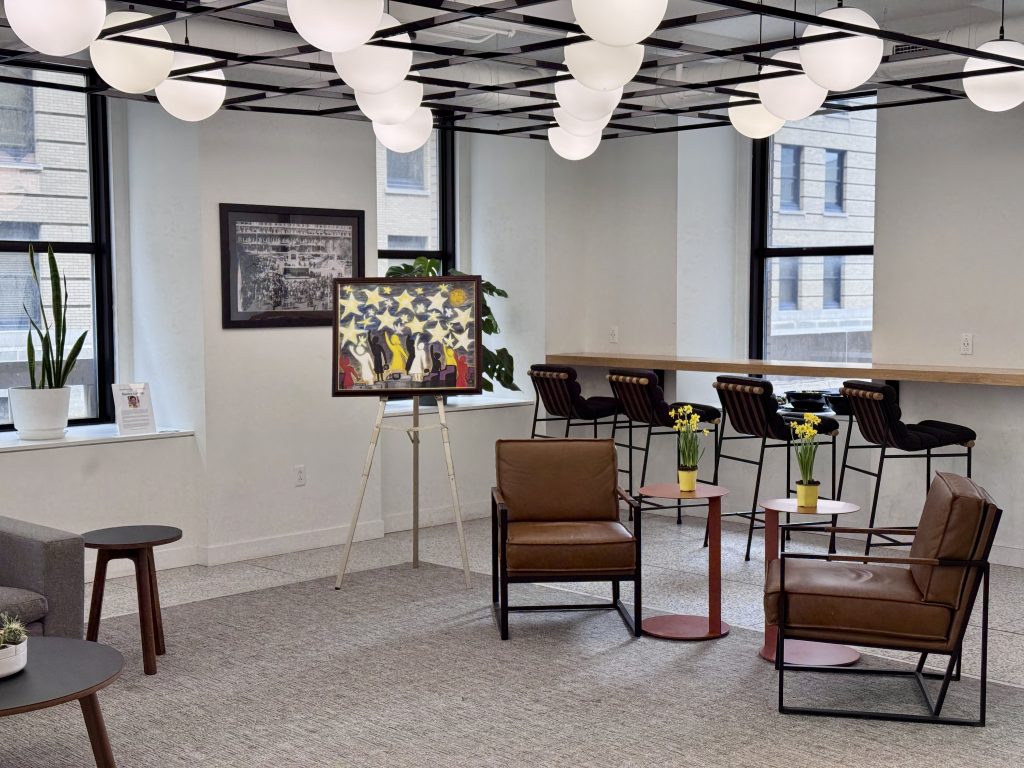 A modern office lounge with brown leather chairs, small tables with yellow flowers, a potted plant, a framed painting on an easel, and large windows letting in natural light. Spherical ceiling lights hang above.