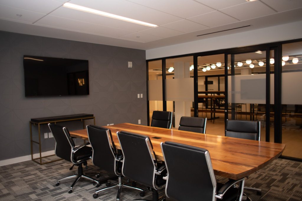 A modern conference room with a wooden table surrounded by black chairs, a large wall-mounted TV, patterned gray carpet, and glass walls revealing an adjacent area with lights.