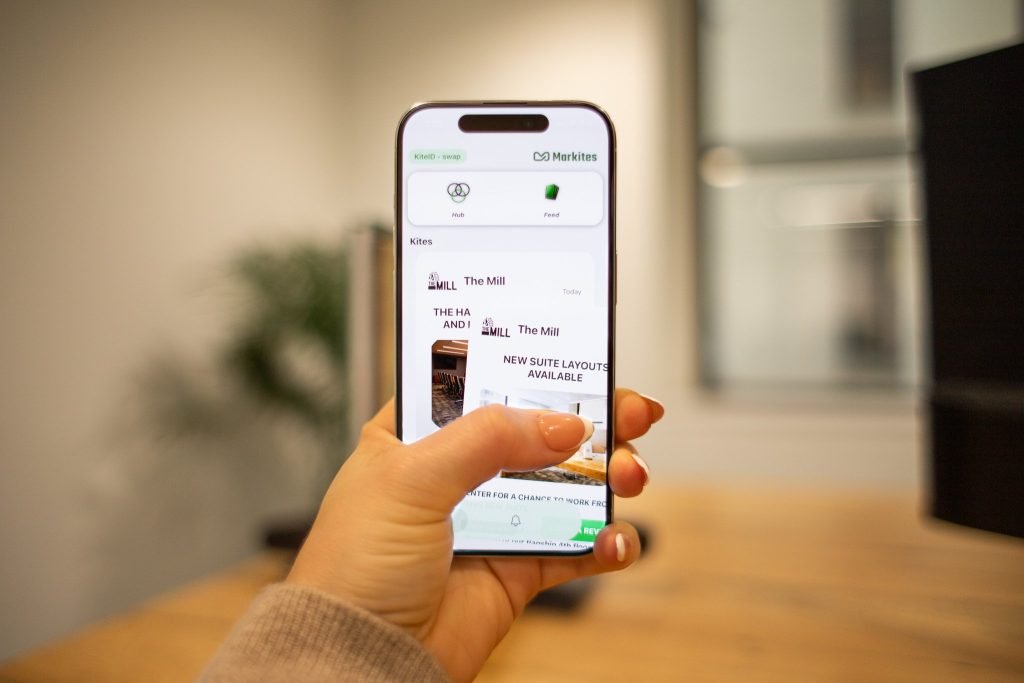 A person holds a smartphone in one hand, browsing an app that displays property listings. The background shows a blurred office setting with a wooden desk and a plant.