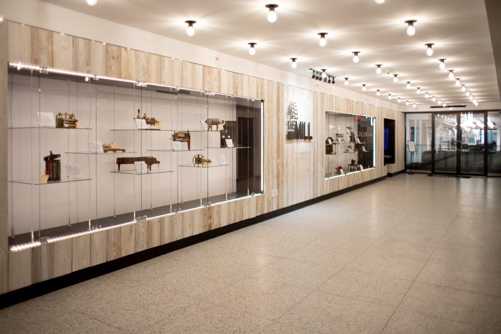 A well-lit hallway with display cases on the left wall showcasing small vintage objects and artifacts, leading to glass doors at the end. The ceiling has rows of round light fixtures.