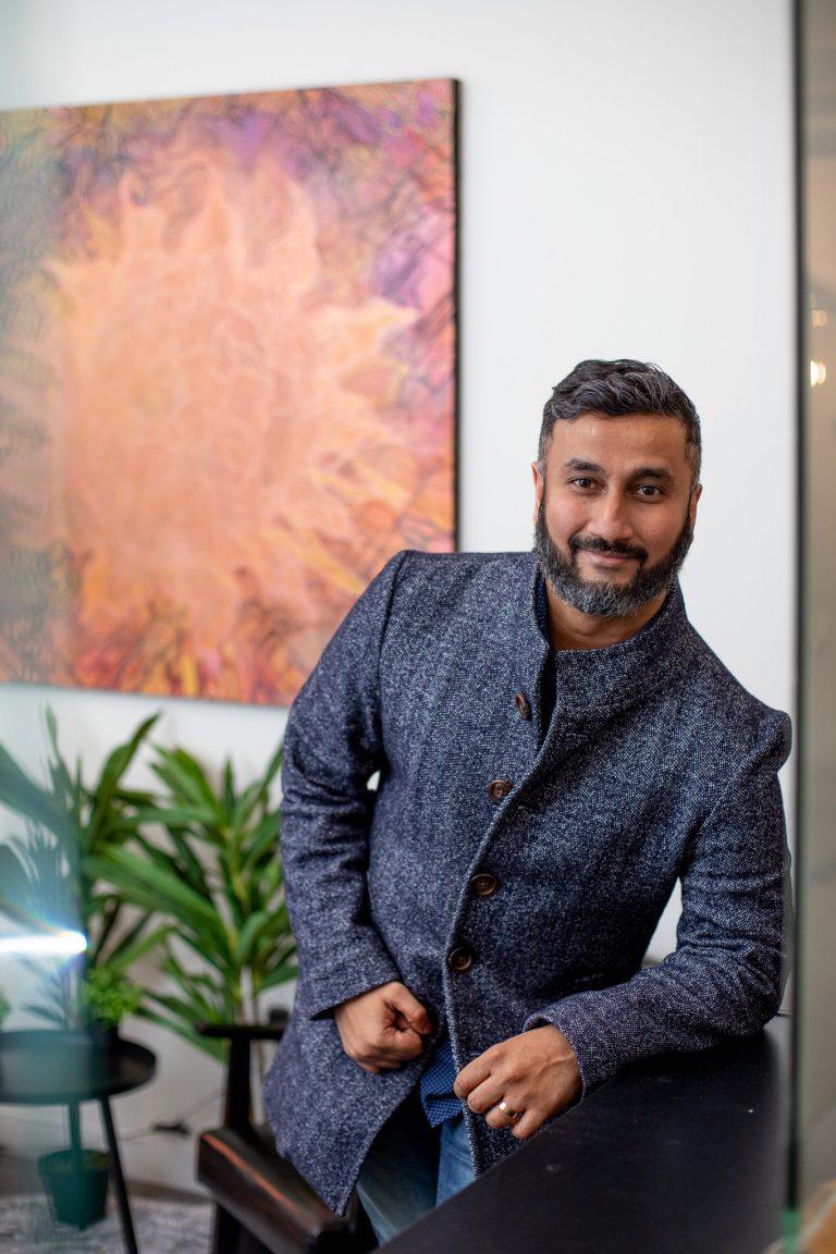 A man with short, dark hair and a beard leans on a table, wearing a buttoned gray jacket. Behind him are green potted plants and a colorful, abstract artwork on a white wall.