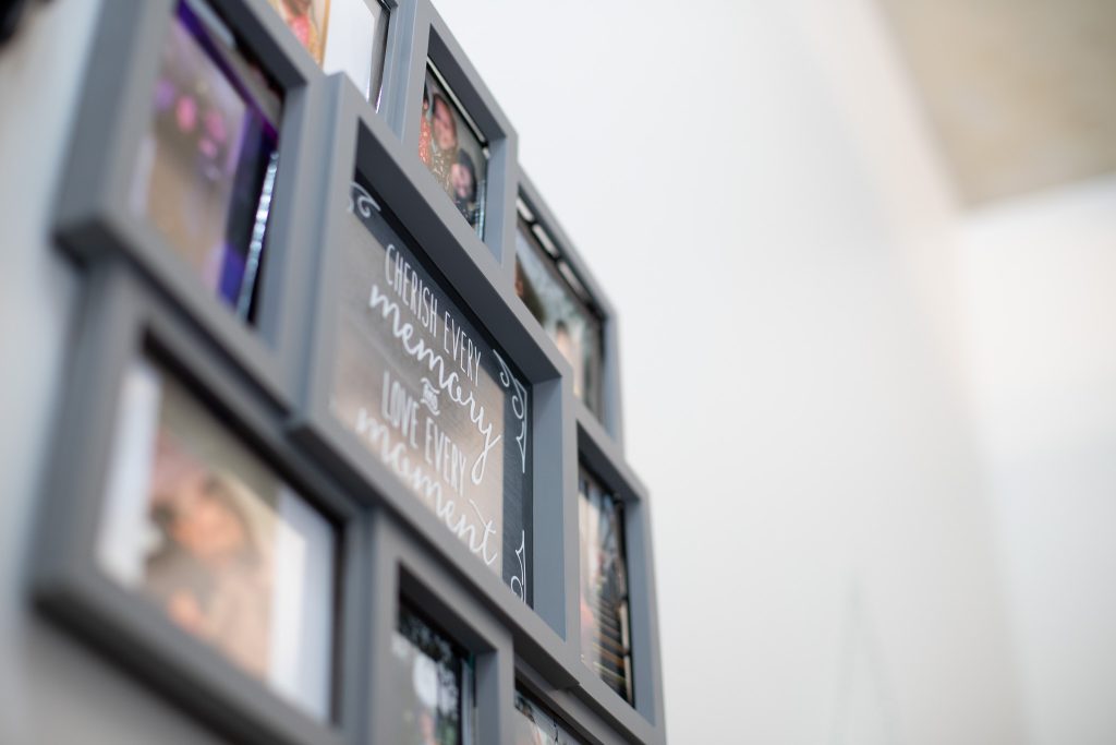 A close-up of a black multi-photo frame on a white wall, featuring several blurred photos and a central sign with cursive text that reads, “Cherish every memory, love every moment.”.