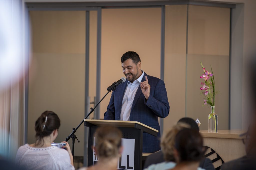 A man in a suit speaks at a podium with a microphone, smiling and gesturing with one finger raised. People are seated in front of him. A vase with pink flowers is on a nearby counter.