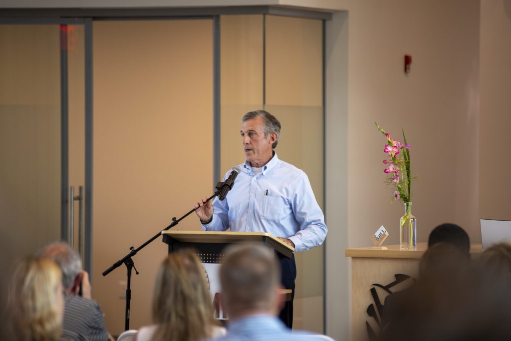 A man speaks at a podium with a microphone in front of an audience. There is a vase with pink flowers on a side table next to him. The room appears to be a conference or meeting space.